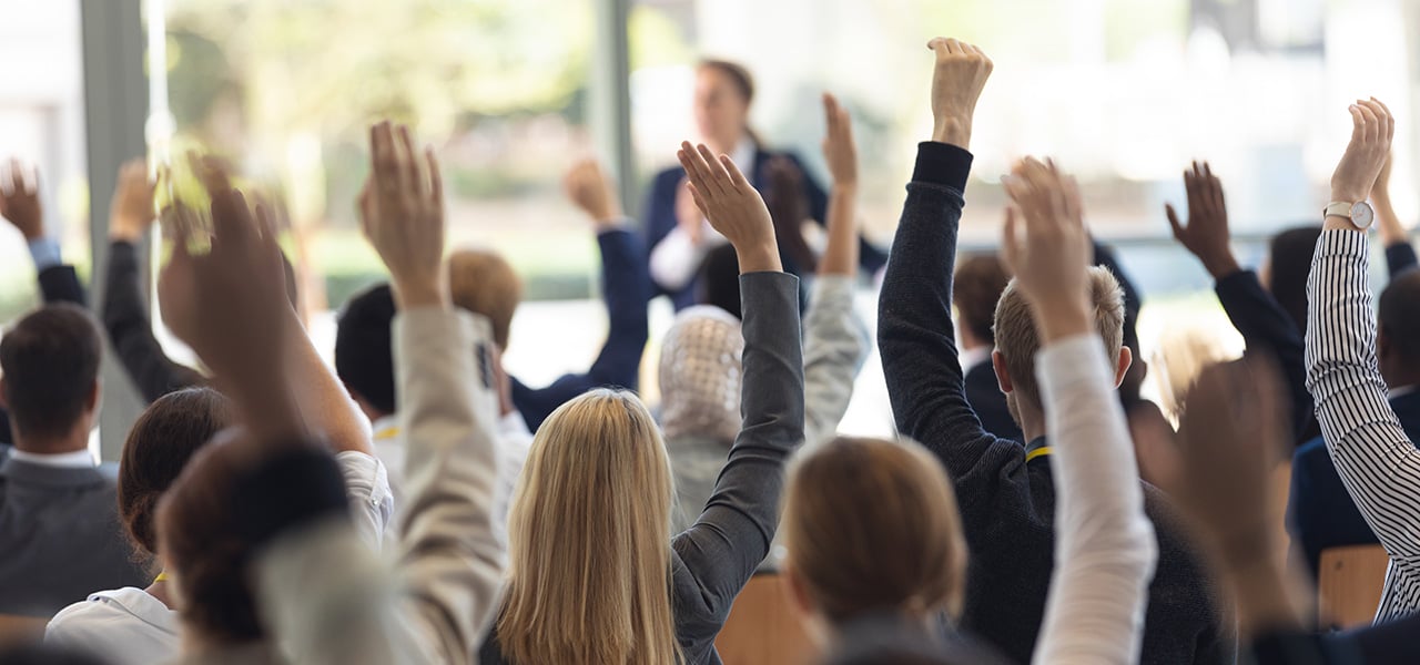konferencetolkning-til-moeder Deltagere rækker hånden op under en konference, mens en person taler i baggrunden – et typisk scenarie hvor konferencetolkning sikrer, at alle deltagere forstår budskabet uanset sprog.